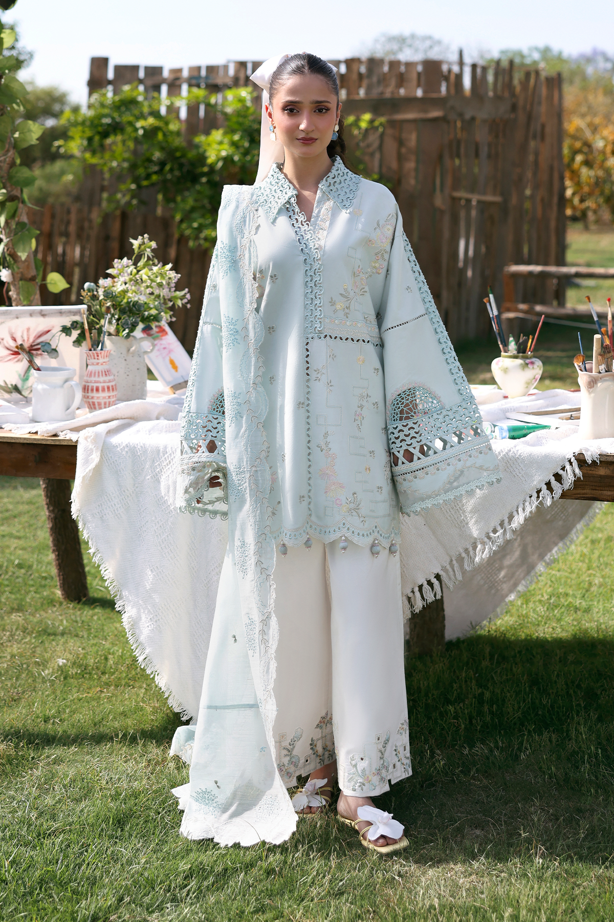 Woman in a light blue embroidered outfit standing outdoors with a table and plants in the background
