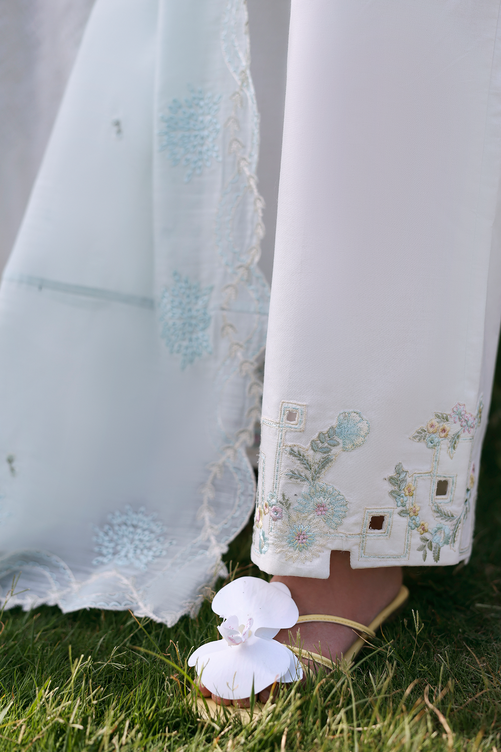 White embroidered pants with floral patterns and a white sandal on grass