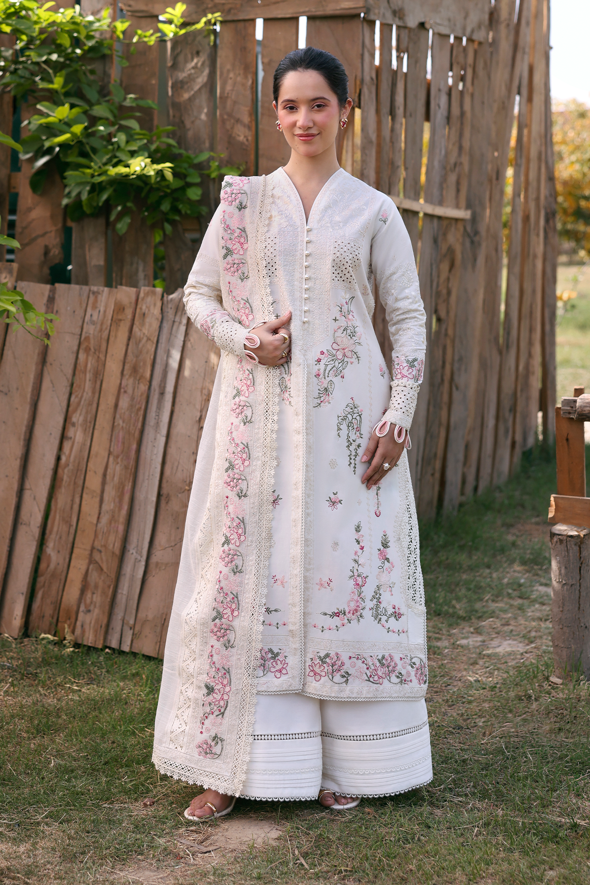 Woman wearing a white embroidered traditional outfit standing outdoors with a wooden fence and greenery in the background.