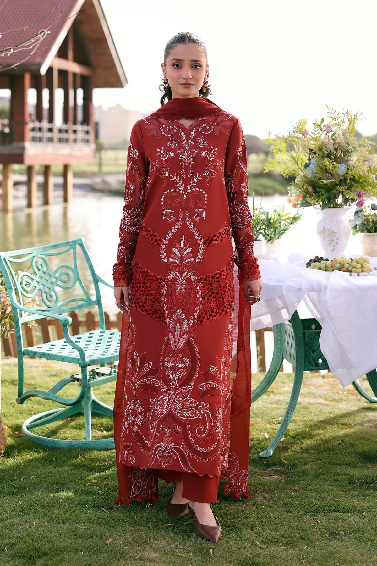 Woman in a red dress with white patterns standing outdoors near a table with flowers and chairs.