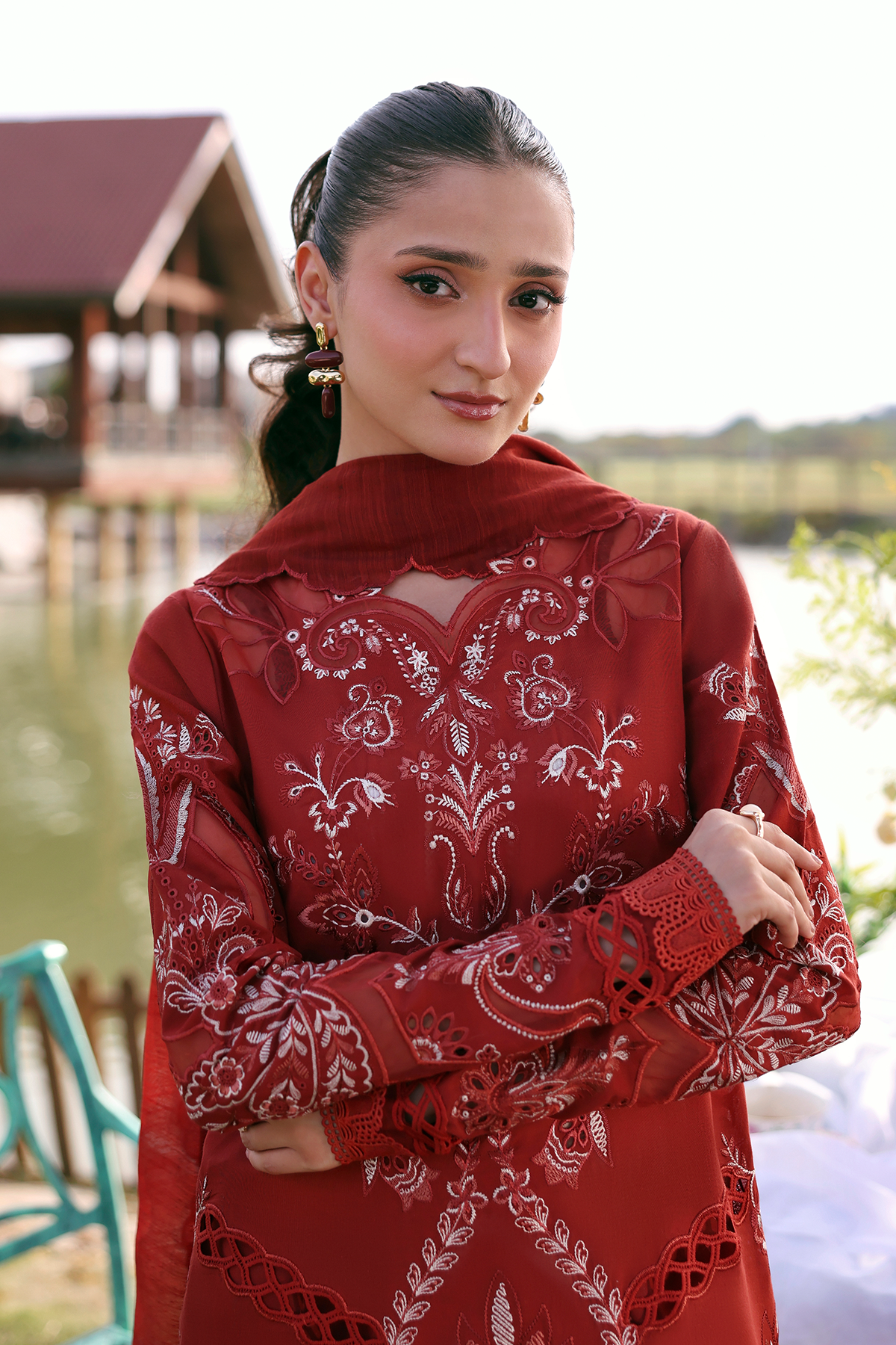 Woman wearing a red traditional outfit with white patterns outdoors by a body of water.