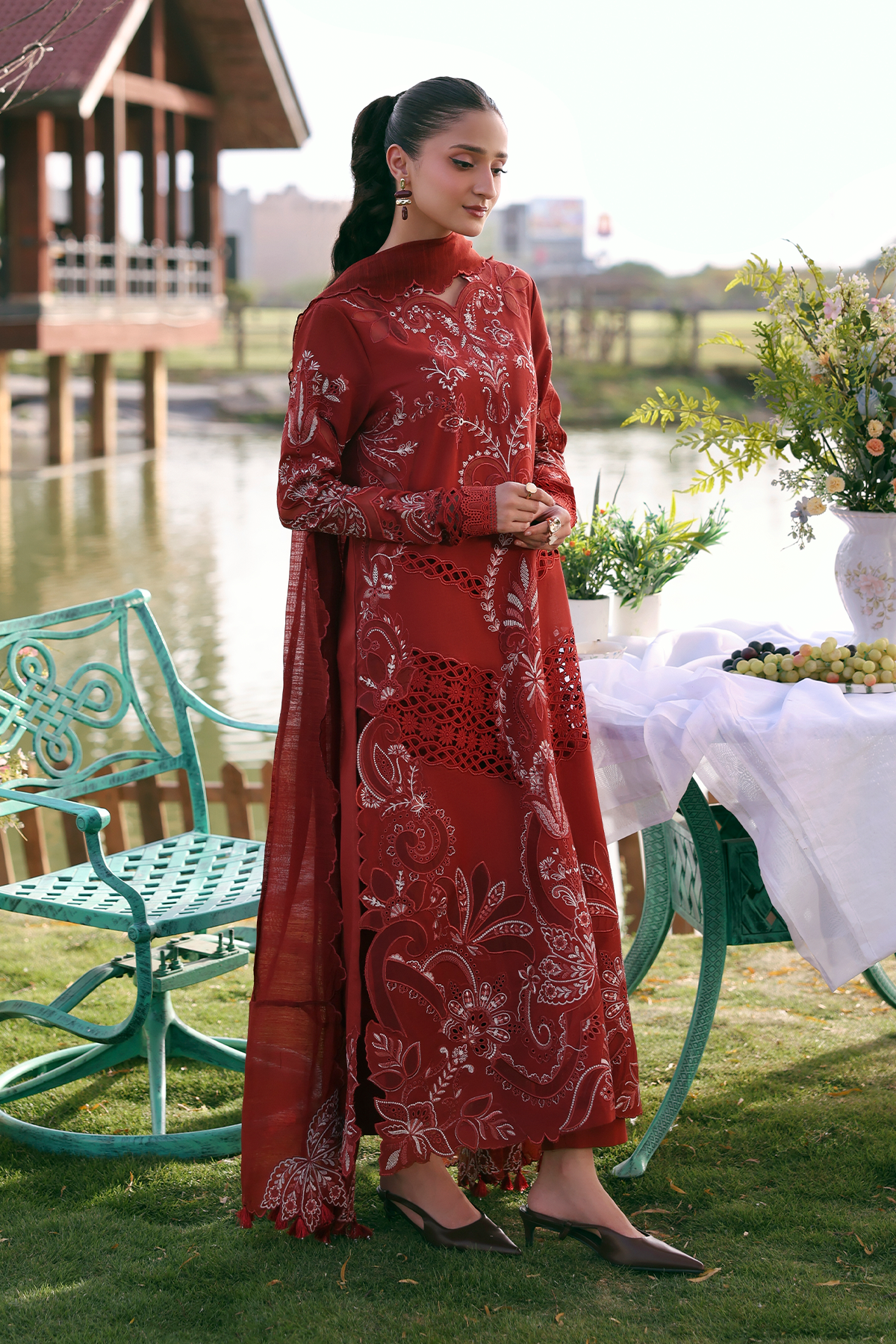 Woman in a red traditional outfit standing outdoors by a lake with greenery and a wooden structure in the background.