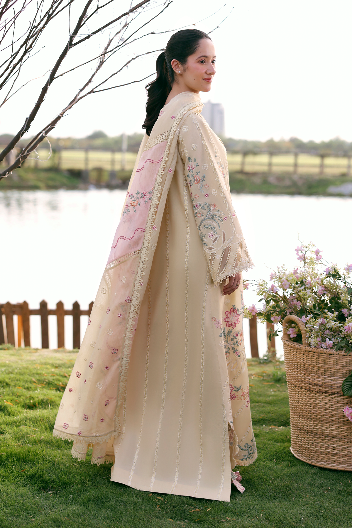 Woman in a traditional outfit standing outdoors by a lake with flowers and a basket.