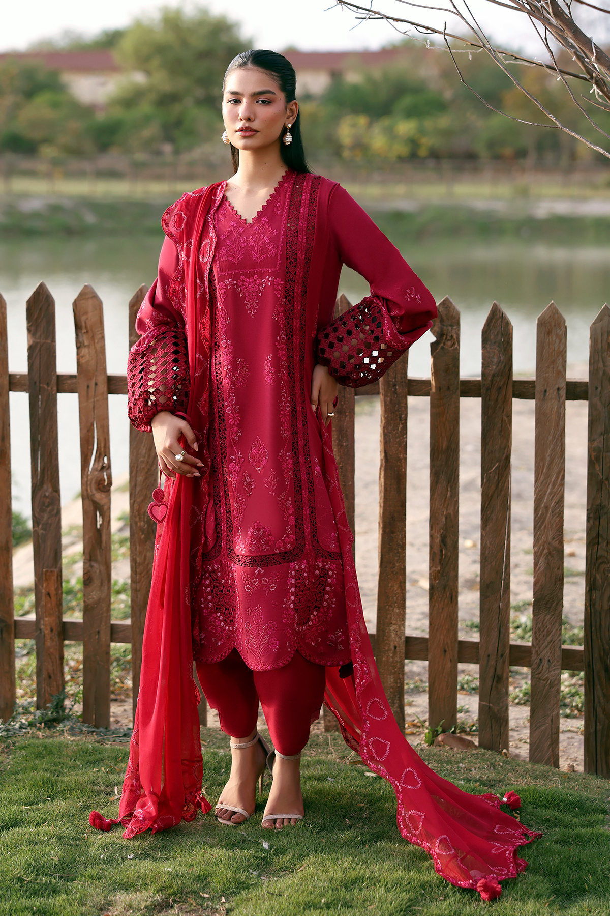 Woman in a red traditional outfit standing outdoors by a wooden fence with a lake in the background.