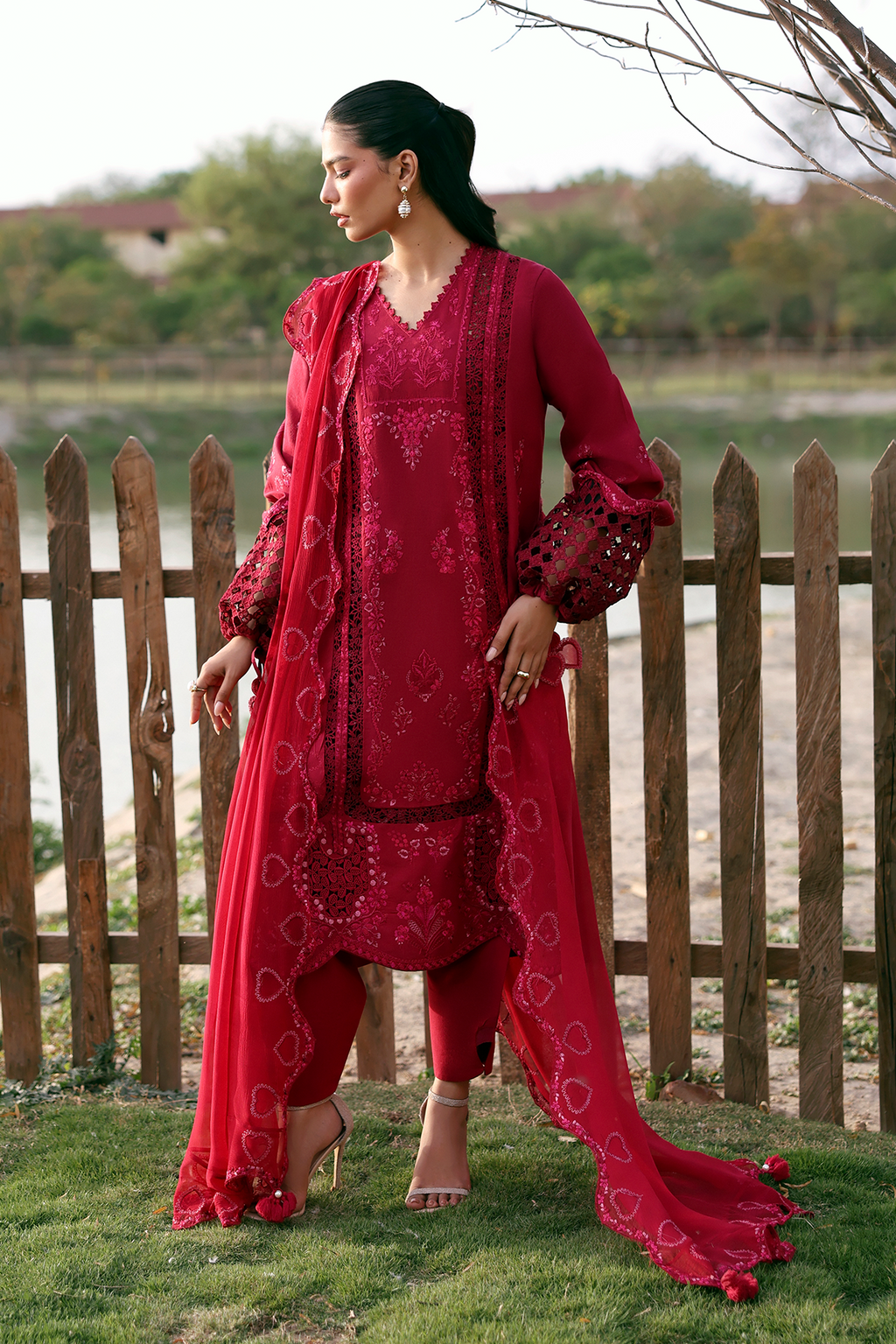 Woman in a red traditional outfit standing outdoors by a wooden fence.