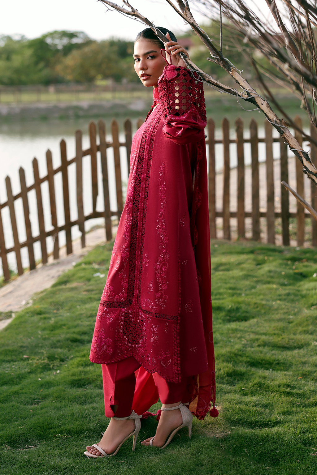 Woman in a red traditional outfit standing outdoors by a wooden fence and body of water.