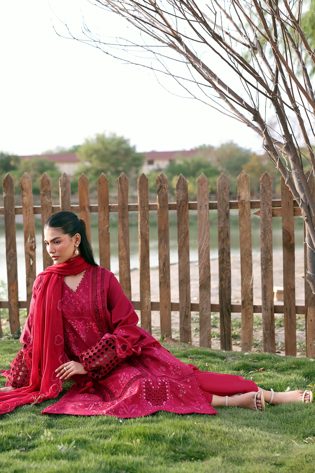 Woman in a red traditional outfit sitting on grass with a wooden fence and trees in the background