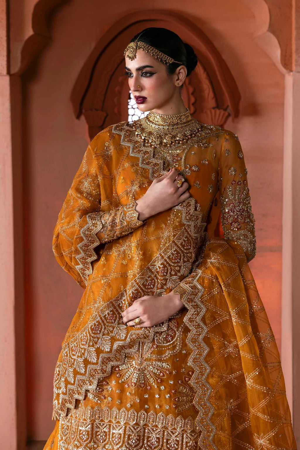 Woman in an orange and gold embroidered traditional outfit with jewelry against a pink archway.