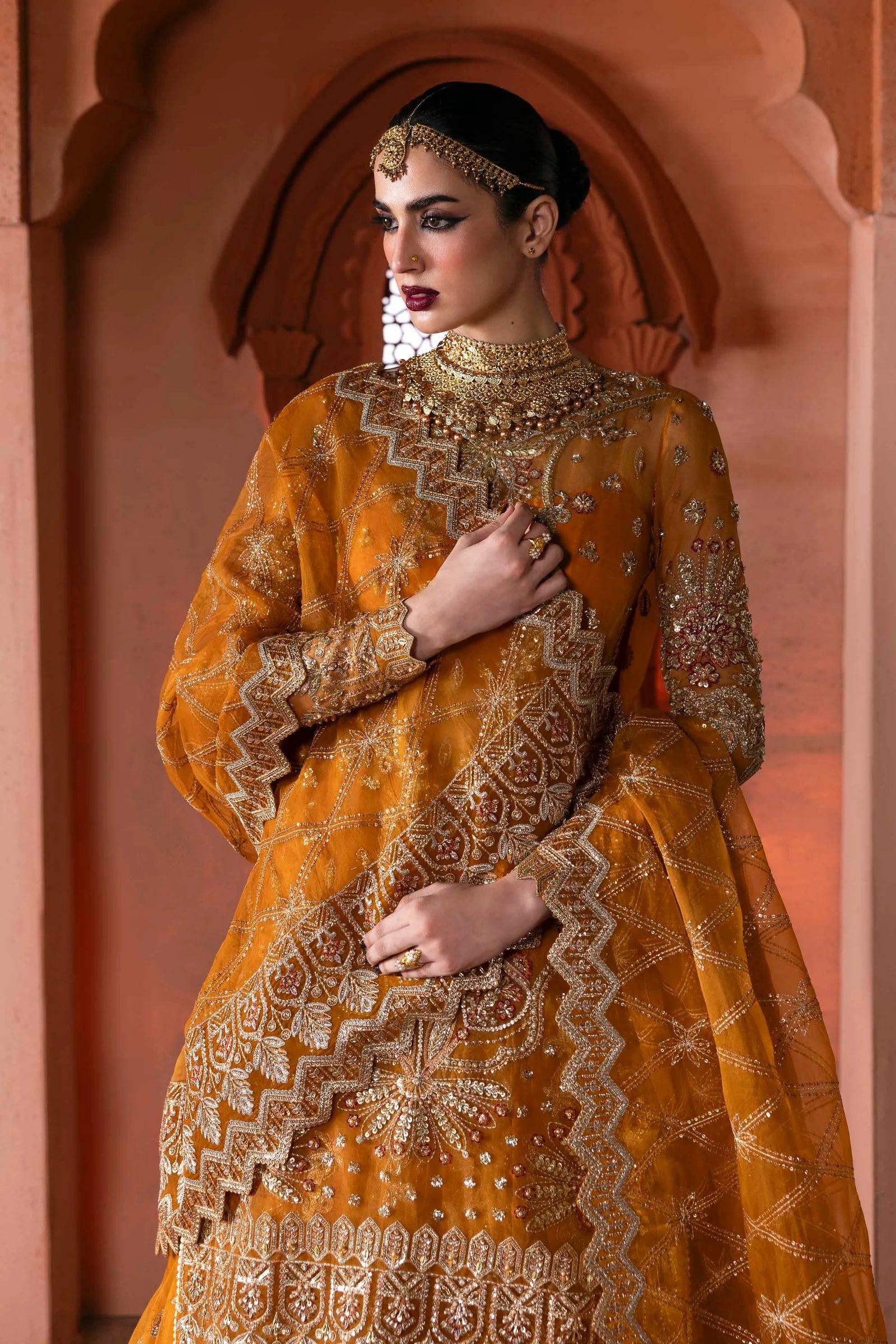 Woman in an orange and gold embroidered traditional outfit with jewelry against a pink archway.