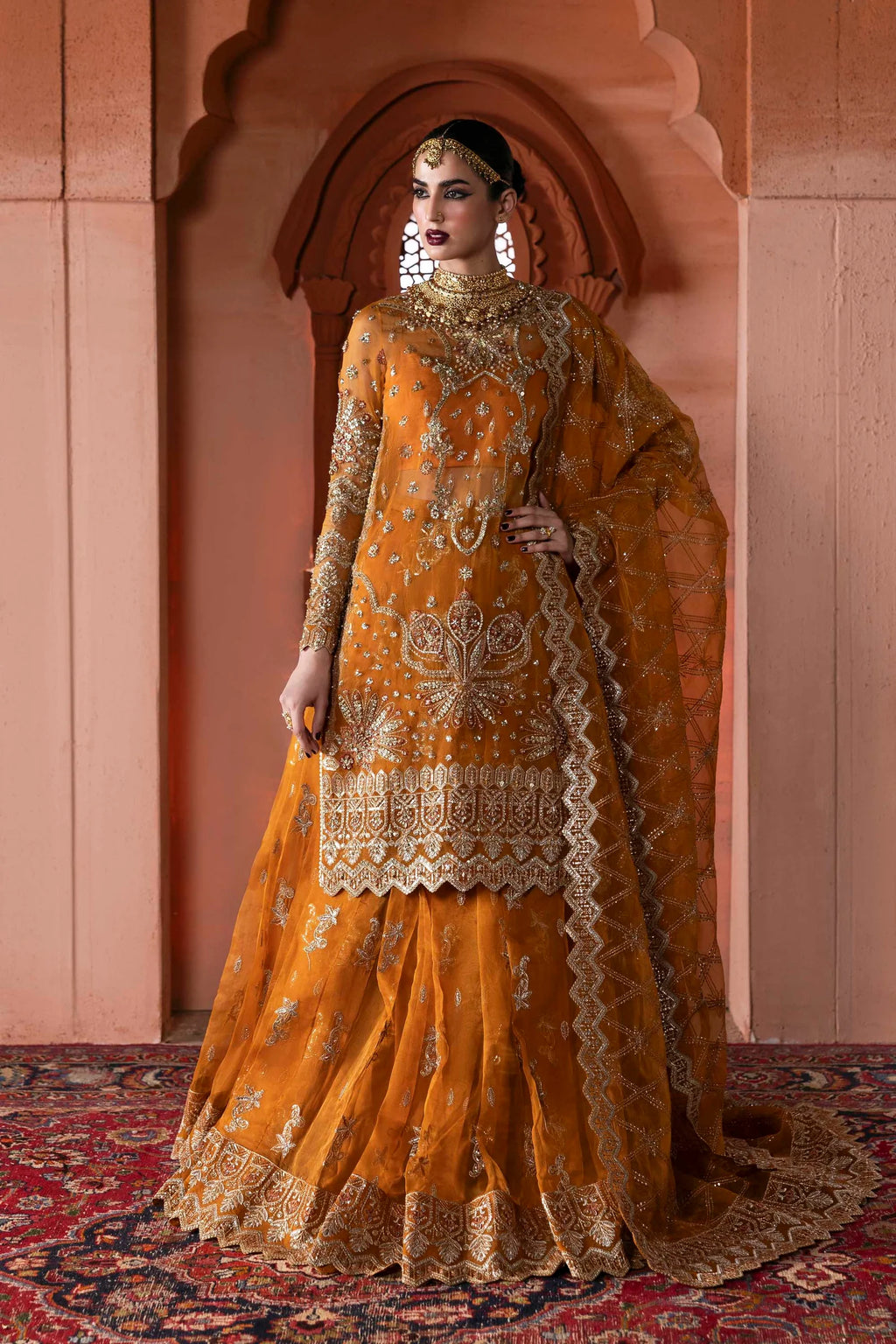 A woman models an ornate traditional Indian outfit in a warm mustard yellow or orange shade with extensive gold embroidery. The outfit consists of a heavily embellished kurta, a matching flared skirt, and a draped dupatta, set against a warm terracotta archway background.