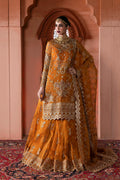 A woman models an ornate traditional Indian outfit in a warm mustard yellow or orange shade with extensive gold embroidery. The outfit consists of a heavily embellished kurta, a matching flared skirt, and a draped dupatta, set against a warm terracotta archway background.
