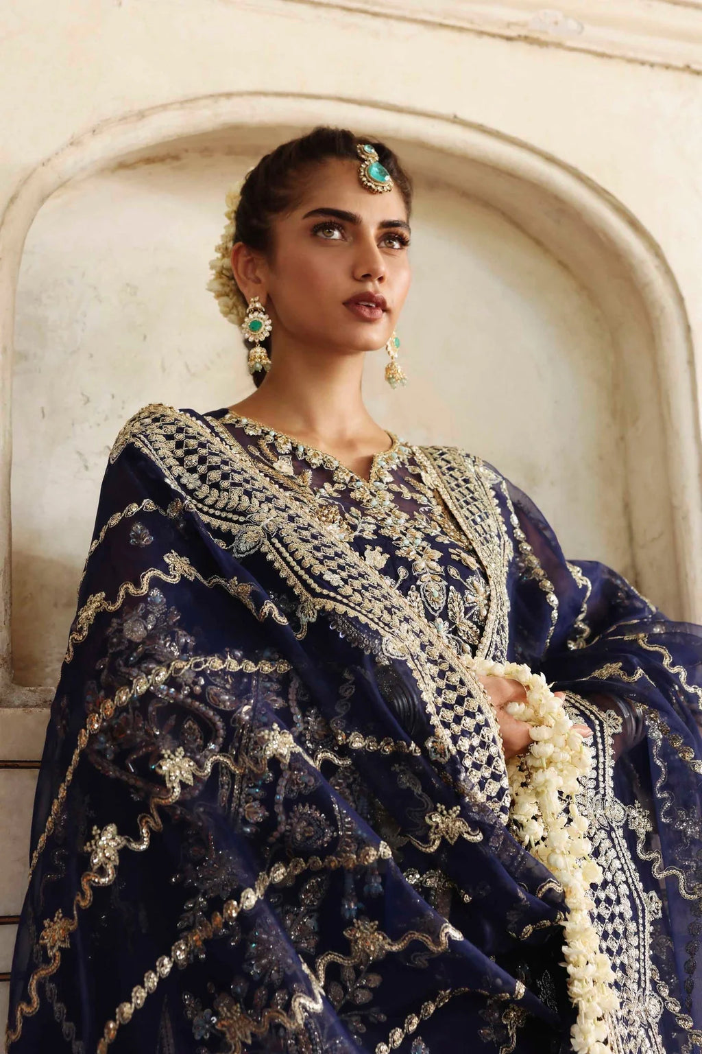 Woman wearing a traditional blue and gold embroidered outfit with jewelry, standing against a neutral wall.