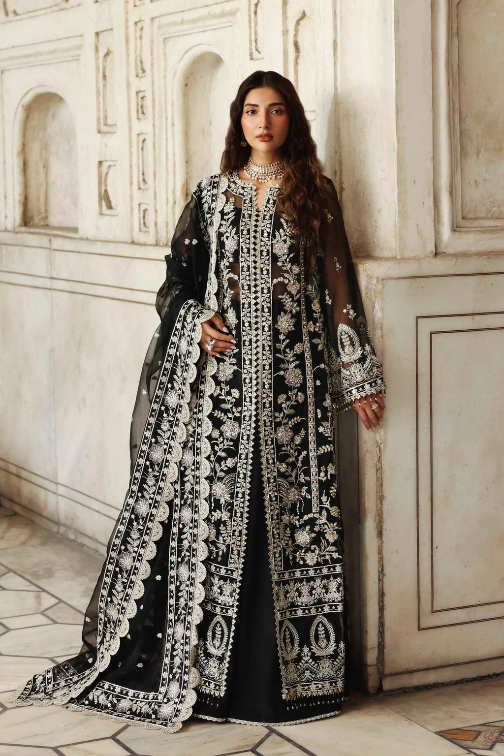 Woman in a black and white embroidered traditional outfit standing against a white marble wall.