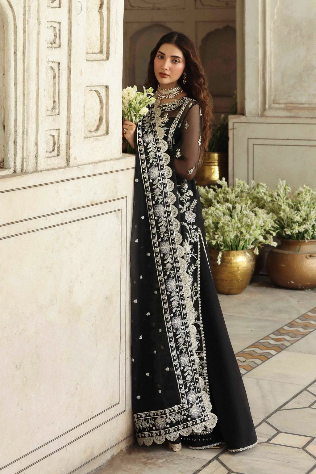 Woman in a black and white embroidered dress standing in a decorative interior setting.
