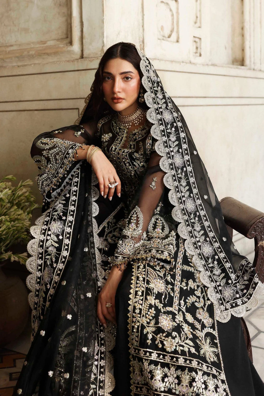 A woman models an ornate black and silver embroidered traditional outfit with a sheer black dupatta featuring intricate silver lace borders, set against a rustic beige stone wall background.