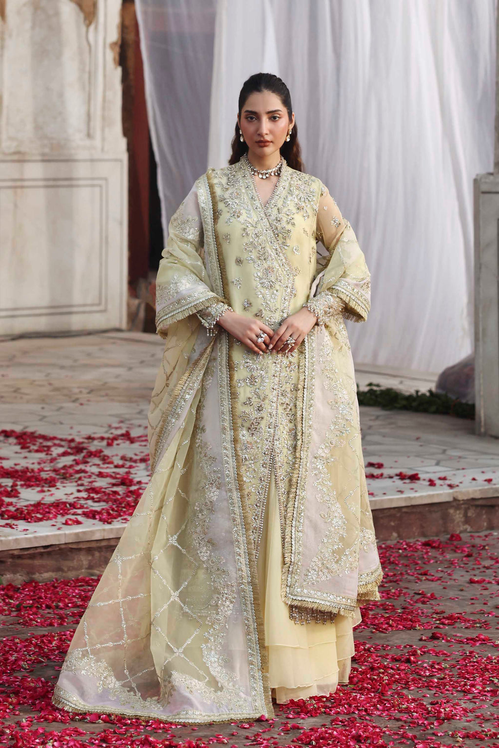 Woman in a traditional outfit standing on a path with rose petals, surrounded by white curtains.