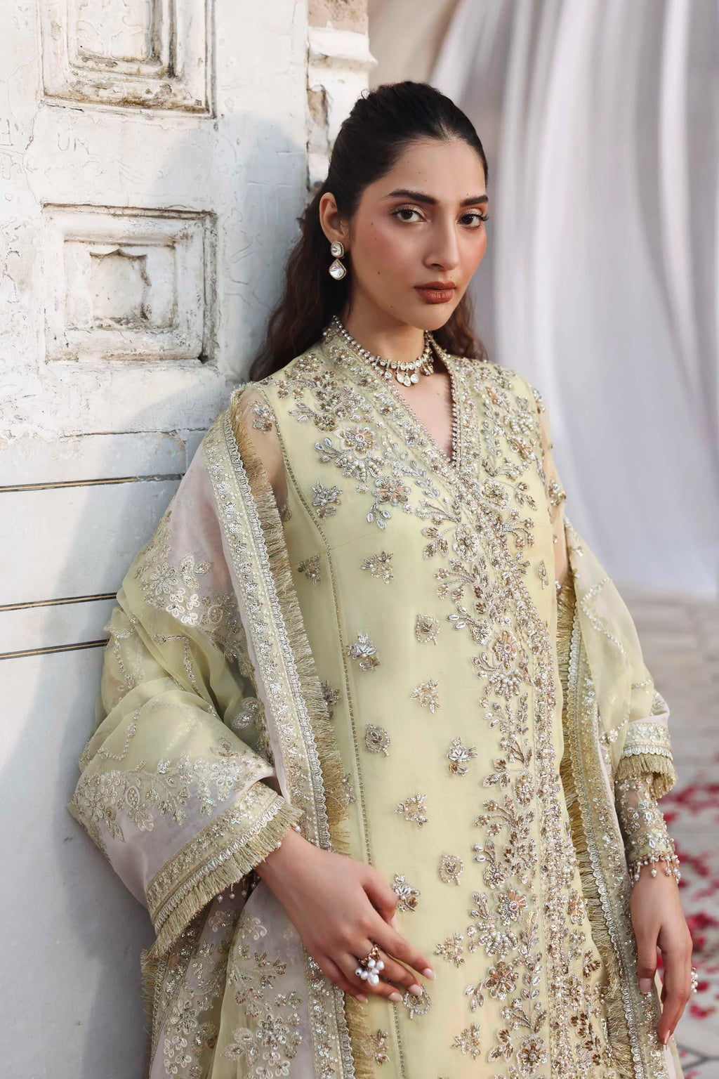 Woman in a light green embroidered traditional outfit standing against a white wall.