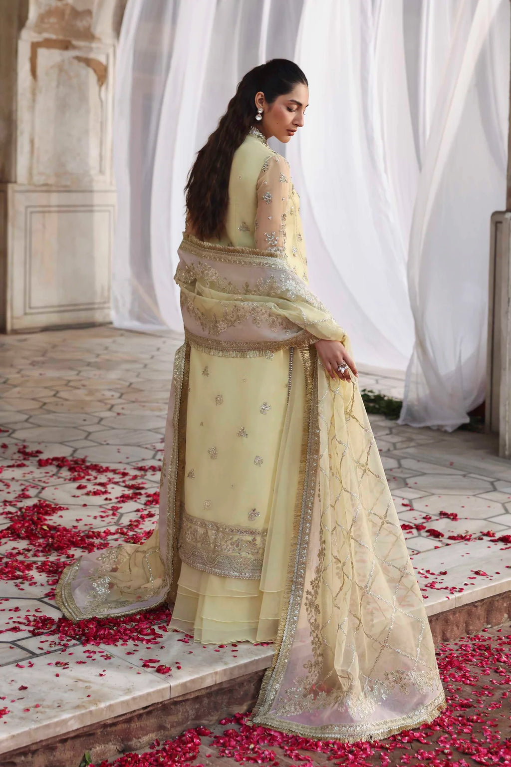 Woman in a traditional outfit standing in a decorated area with white curtains and red flowers on the floor.