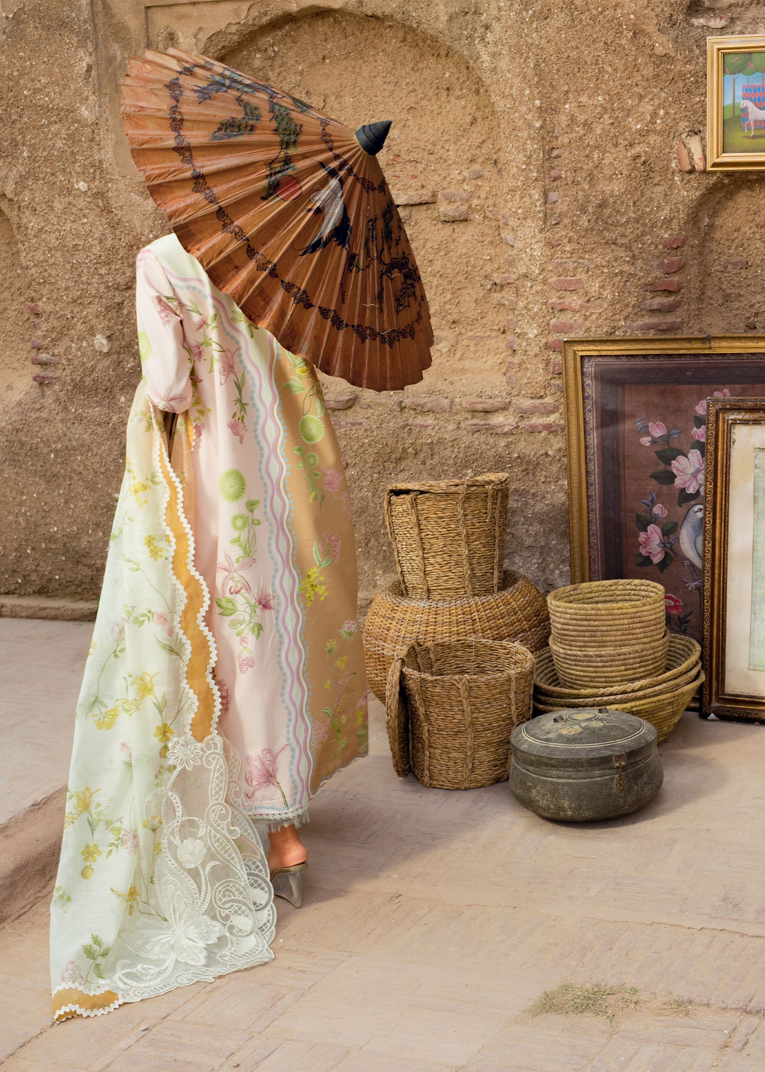 Person in traditional attire with an umbrella standing next to woven baskets and a textured wall.