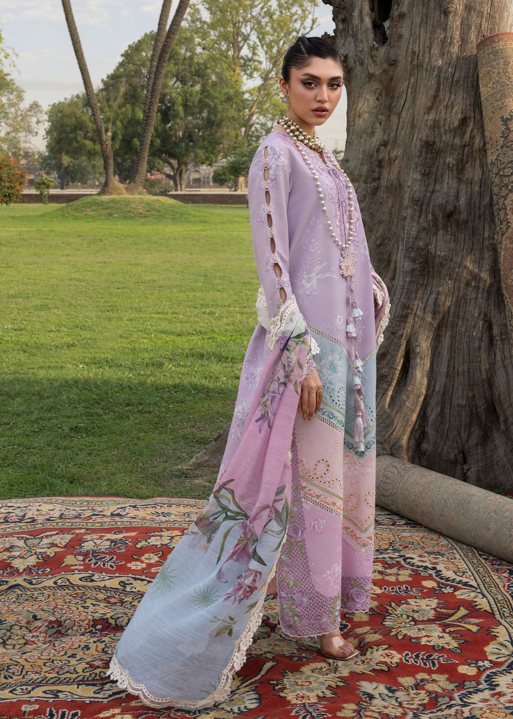 Woman in a light purple floral dress standing outdoors on a rug with trees in the background