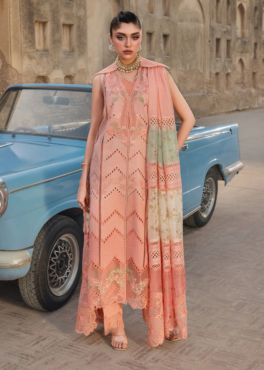 Woman in a peach embroidered dress standing next to a vintage blue car with an old building in the background.