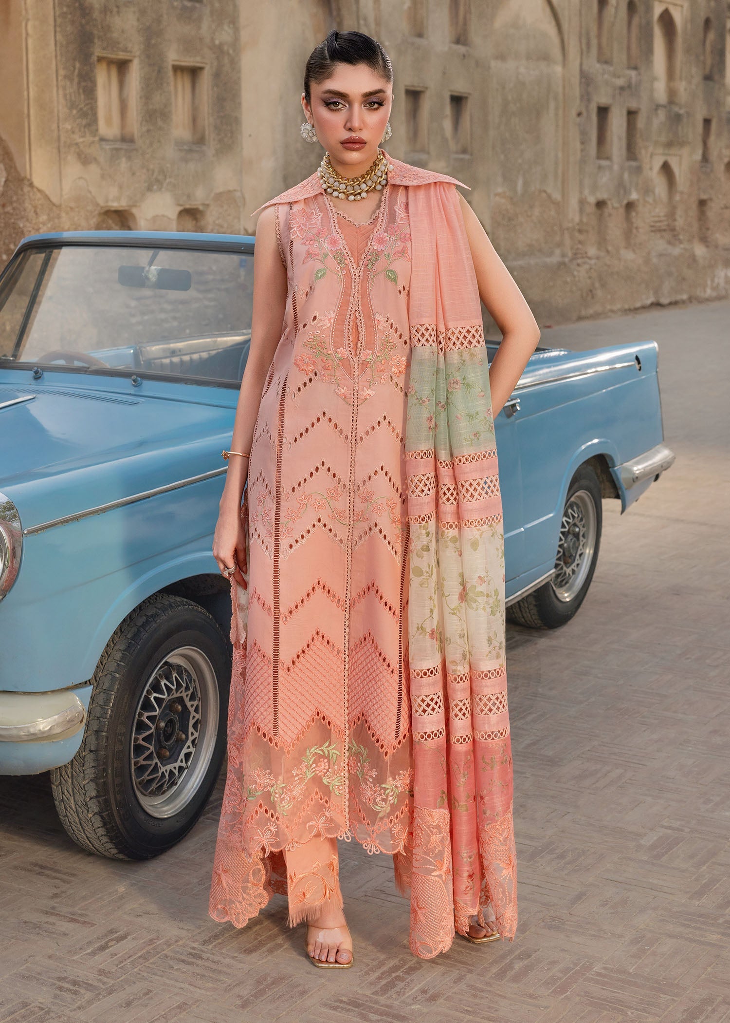Woman in a peach embroidered dress standing next to a vintage blue car with an old building in the background.