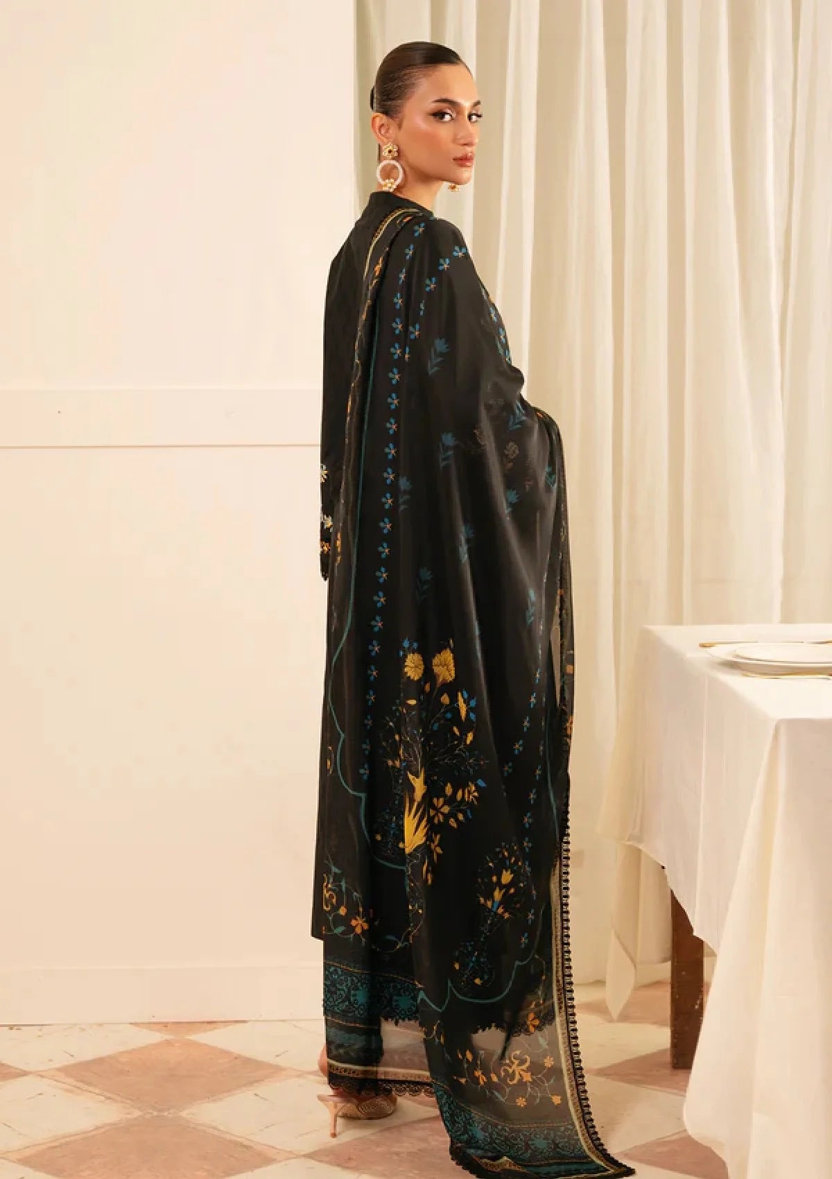 A woman modeling a black traditional outfit with teal and gold floral embroidery, standing in a softly lit indoor setting with cream curtains and a table in the background.