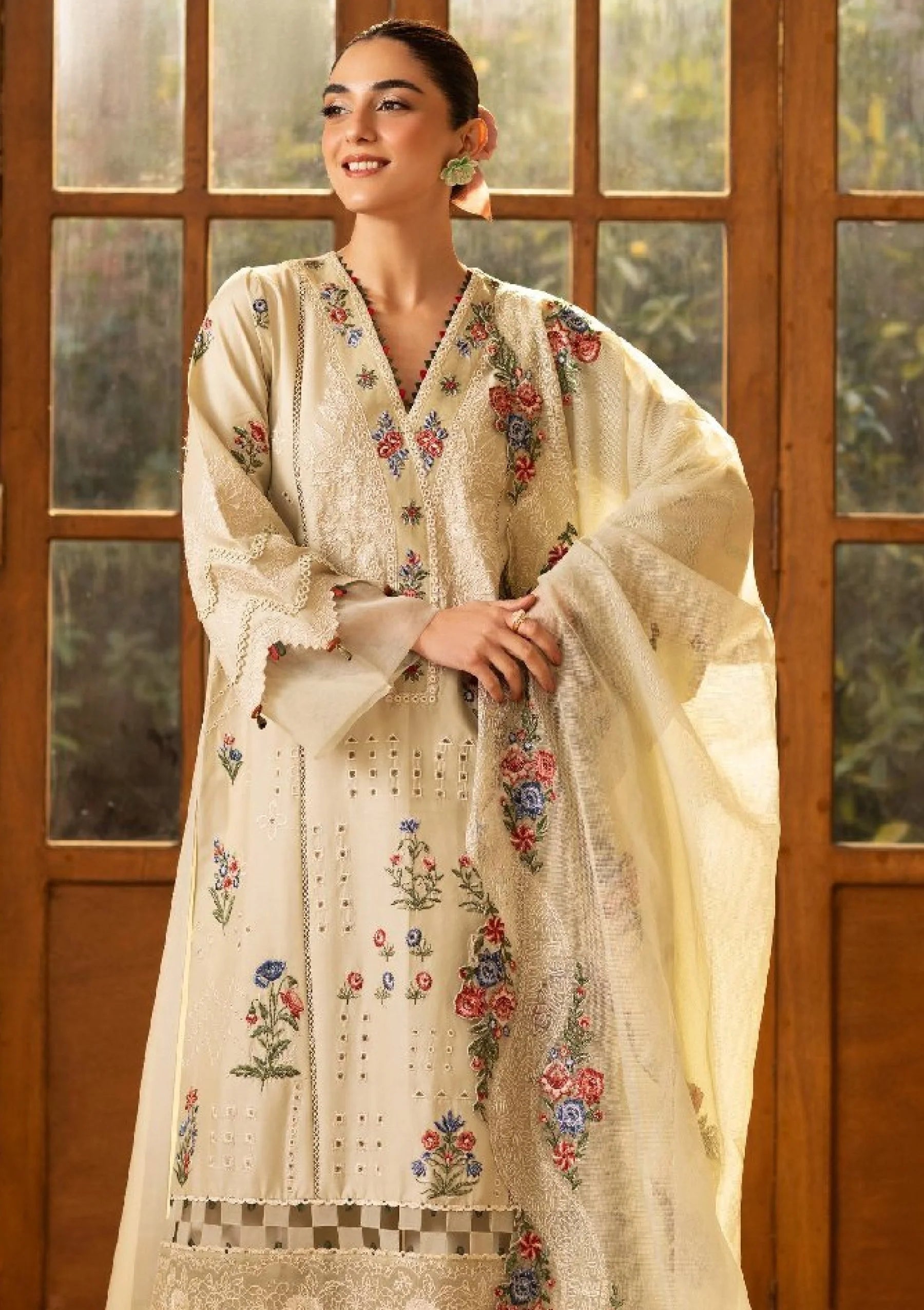 A woman models a traditional cream-colored embroidered kurta set with a matching dupatta, featuring floral and geometric patterns, posed in front of a wooden window frame.