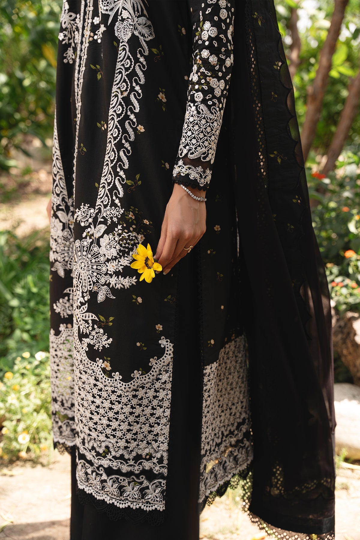Person wearing a black dress with white lace details, holding a yellow flower in a garden setting.