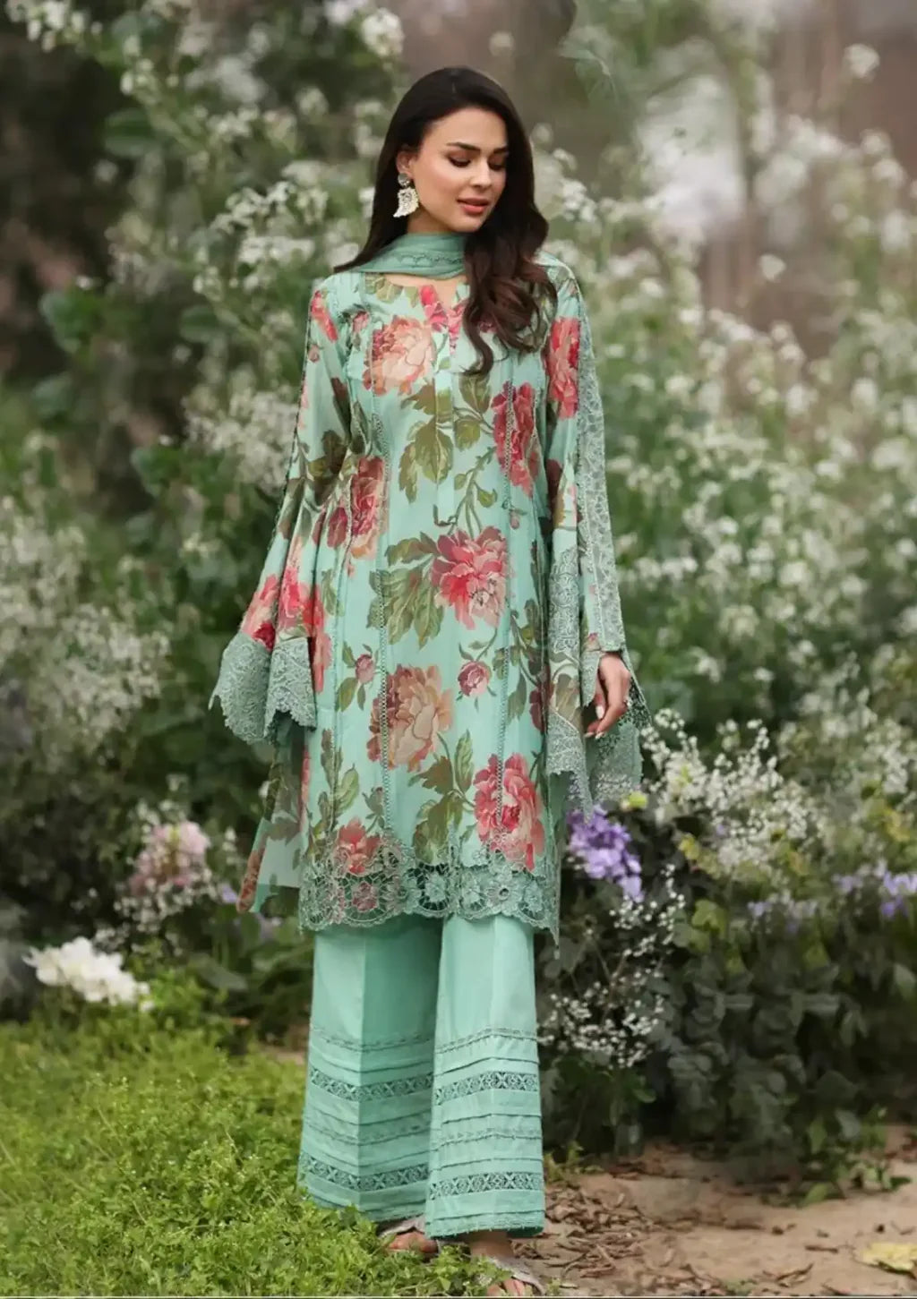 A woman wearing a mint green three-piece traditional outfit with a pink and red floral print, standing in a garden with green grass and white flowers in the background.