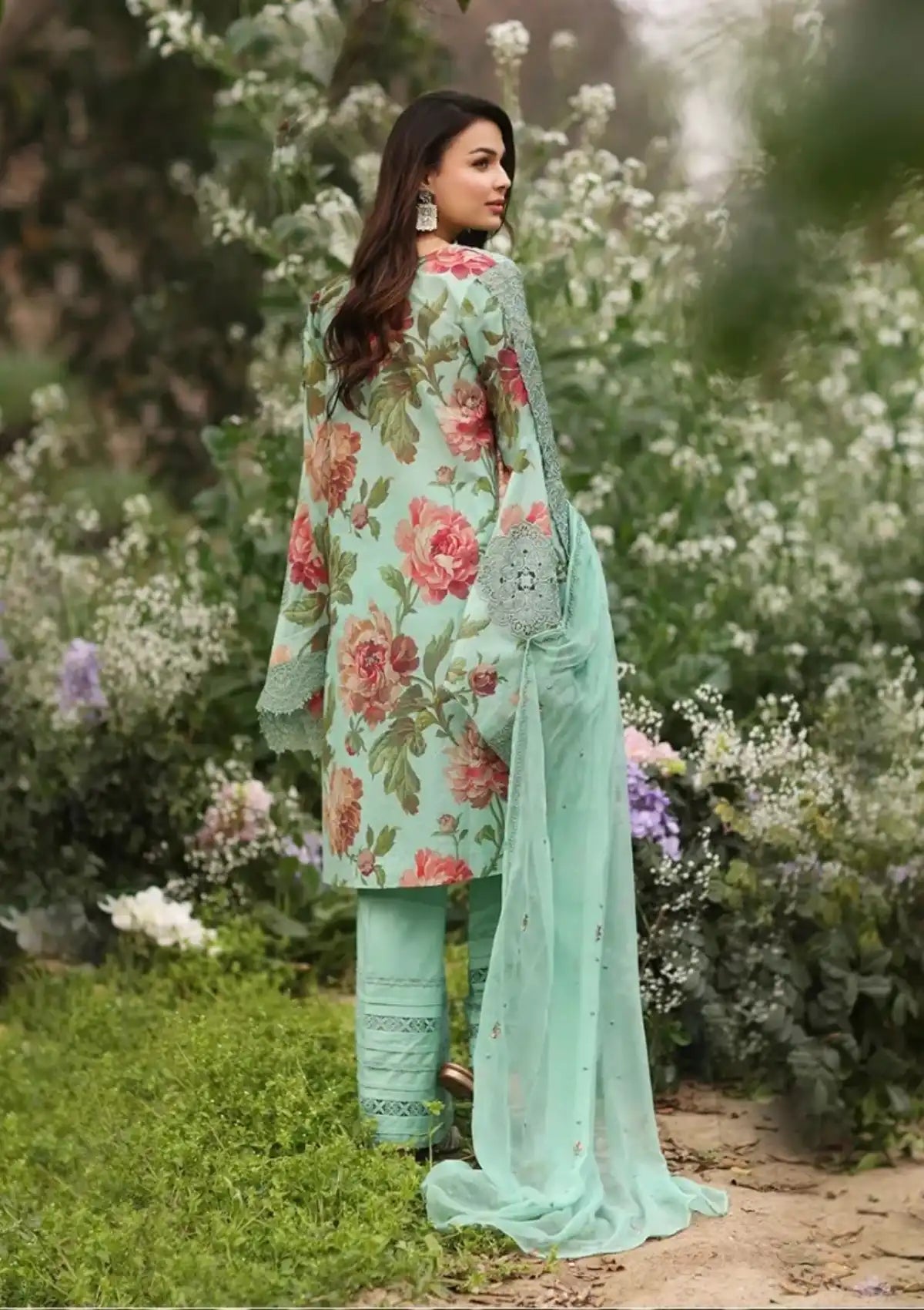 A woman stands outdoors in a garden wearing a mint green traditional outfit with a floral print, matching pants, and a sheer dupatta.