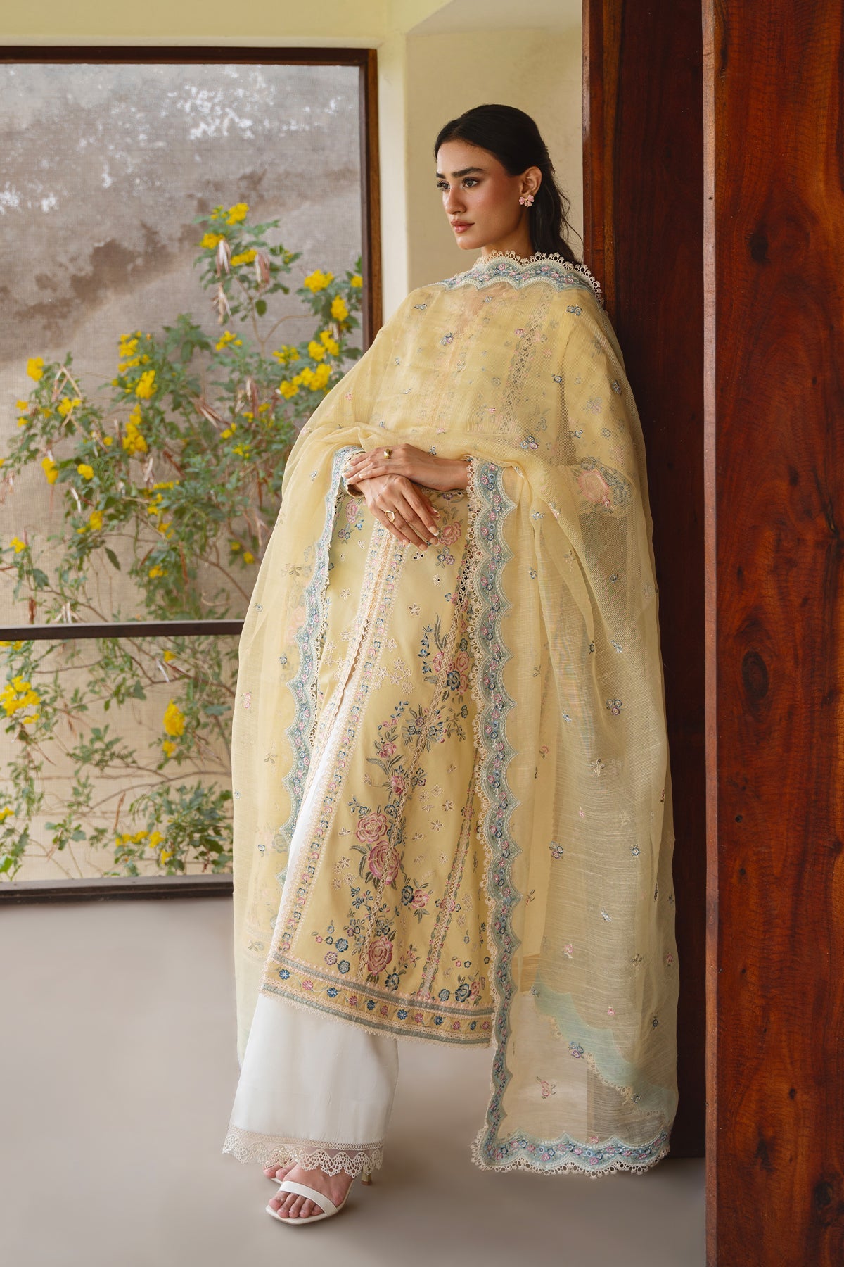 Woman in a light yellow traditional outfit with floral patterns standing indoors.