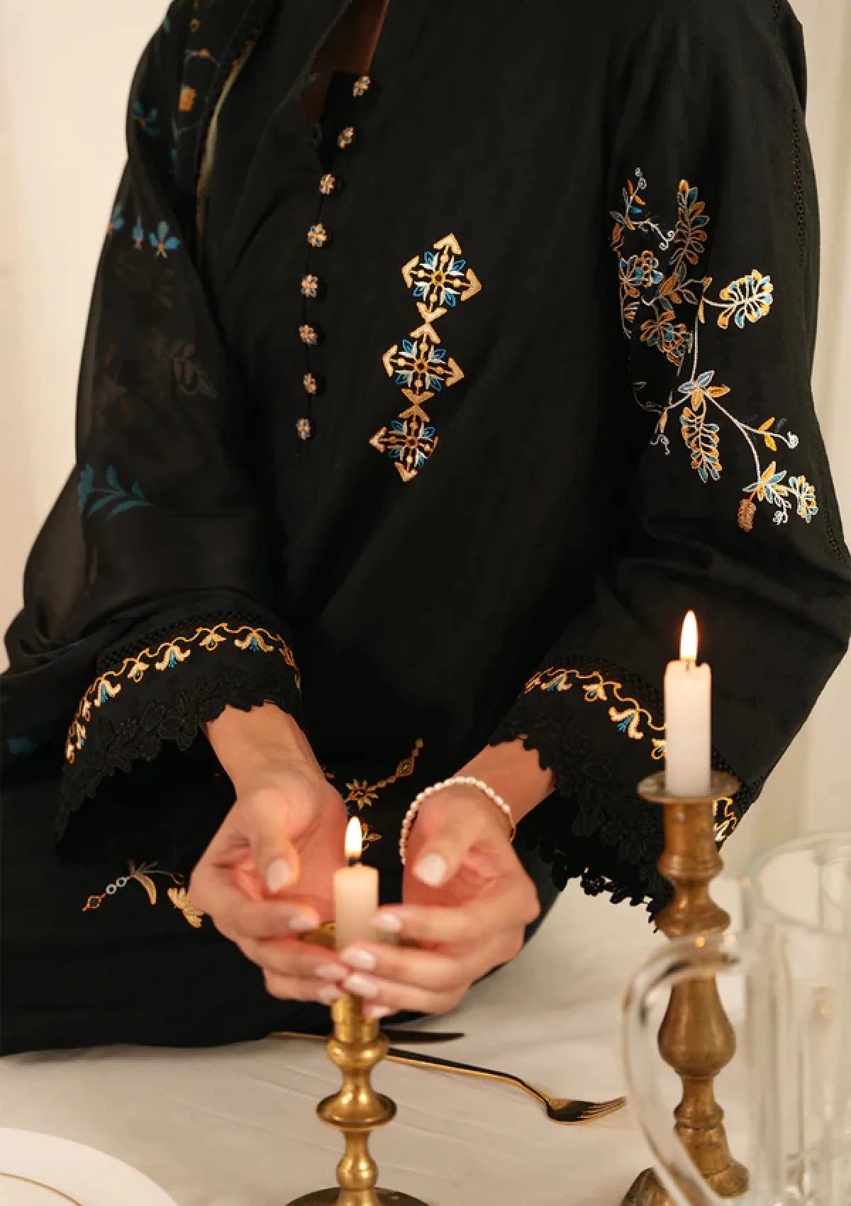 A close-up of a person wearing a black embroidered tunic with gold and teal floral detailing on the sleeves and chest, holding a lit candle in a brass holder at a table with other candles.