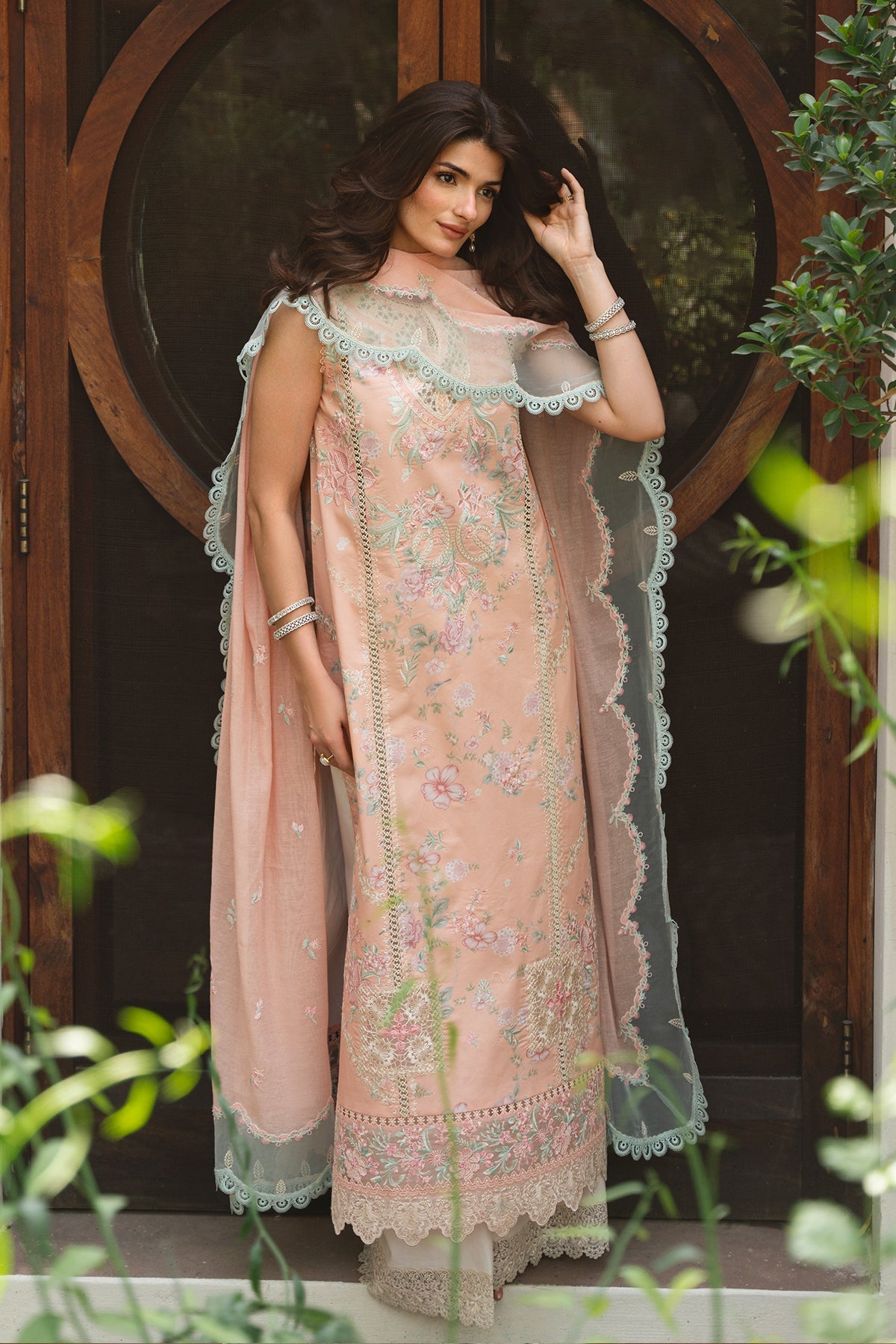 Woman wearing a traditional pink embroidered outfit with a sheer dupatta in front of a wooden door.