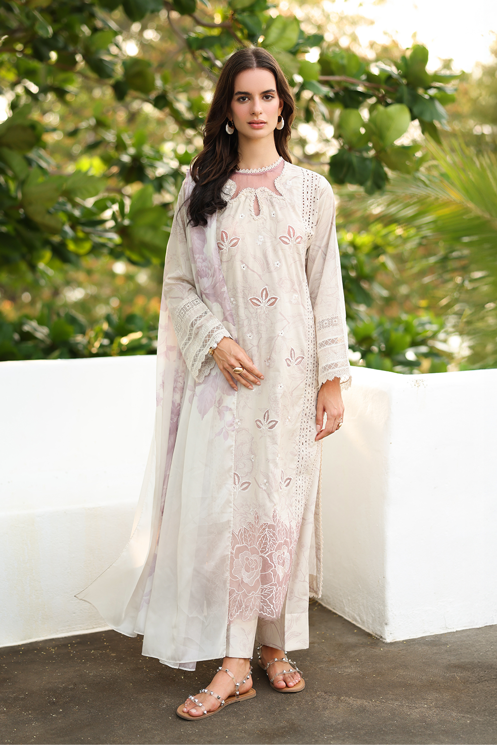 Woman wearing a light-colored embroidered outfit standing outdoors with greenery in the background