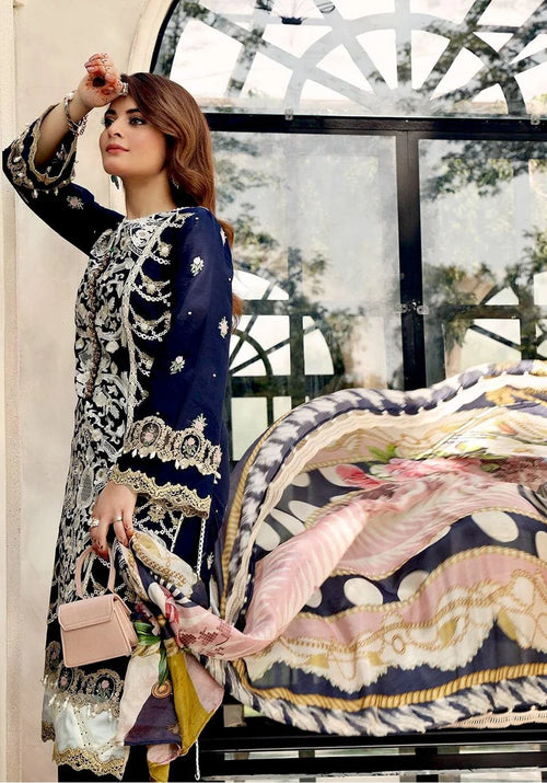 A woman models a navy blue and silver embroidered traditional outfit with a matching patterned scarf and a small light pink handbag, posed in front of a large arched window.