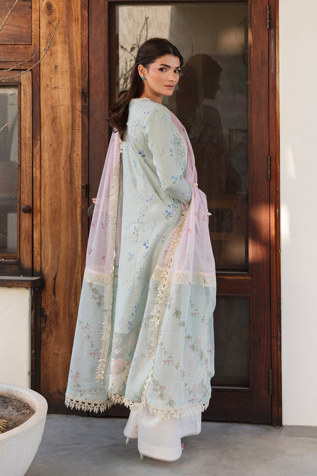 Woman in a light blue traditional outfit with a pink dupatta standing in a room with wooden walls.