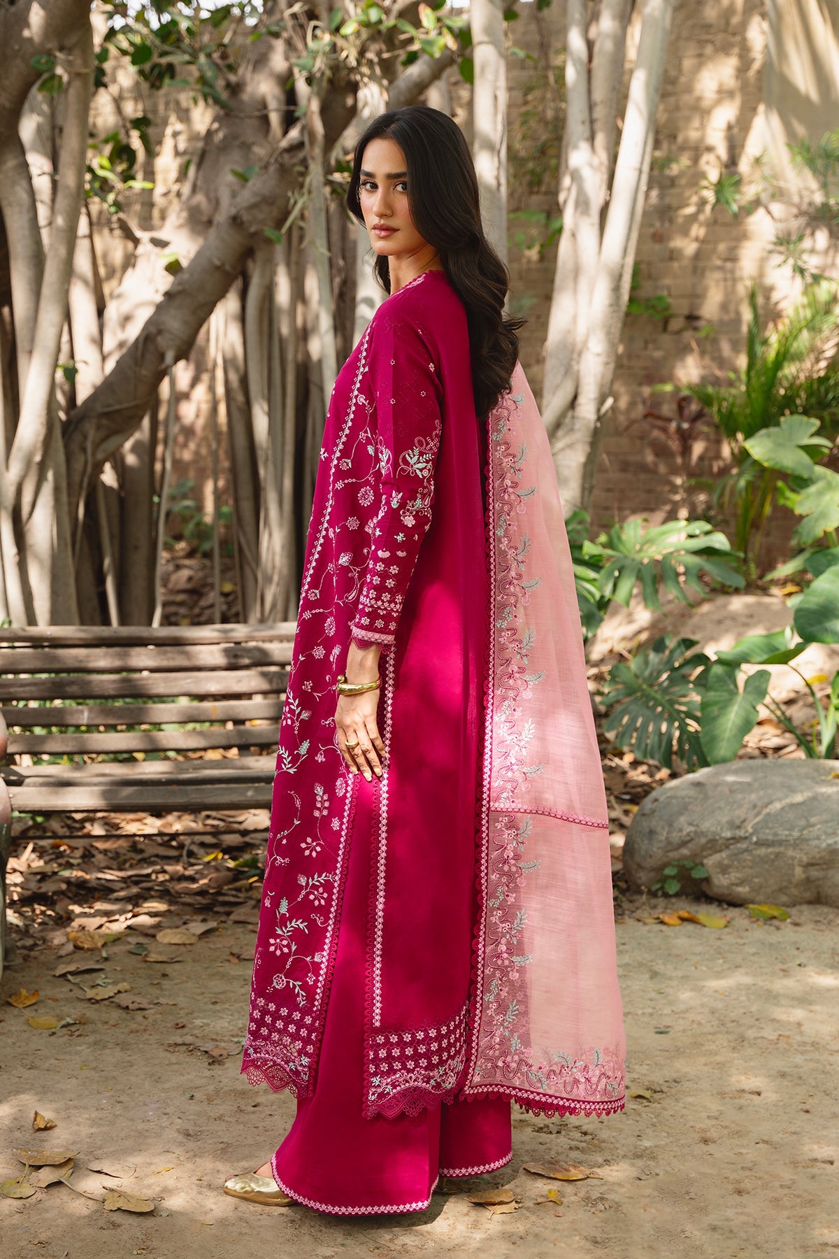 Woman in a pink traditional outfit standing outdoors with trees and a bench in the background