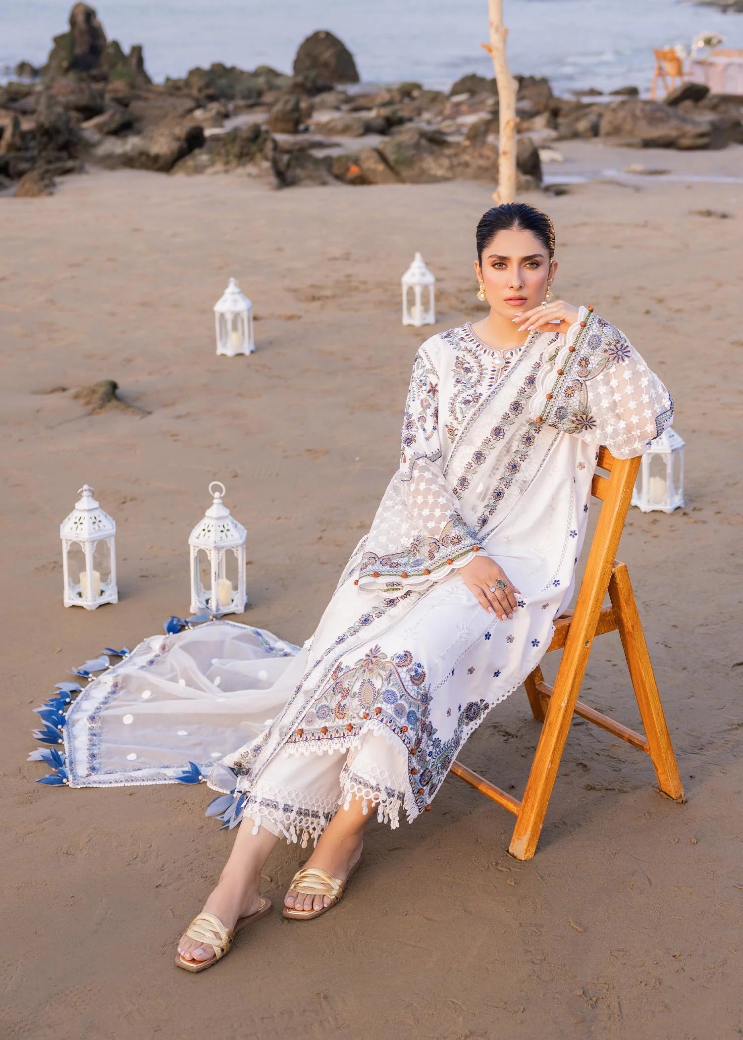 Woman in a white saree sitting on a wooden chair by the beach with lanterns around.