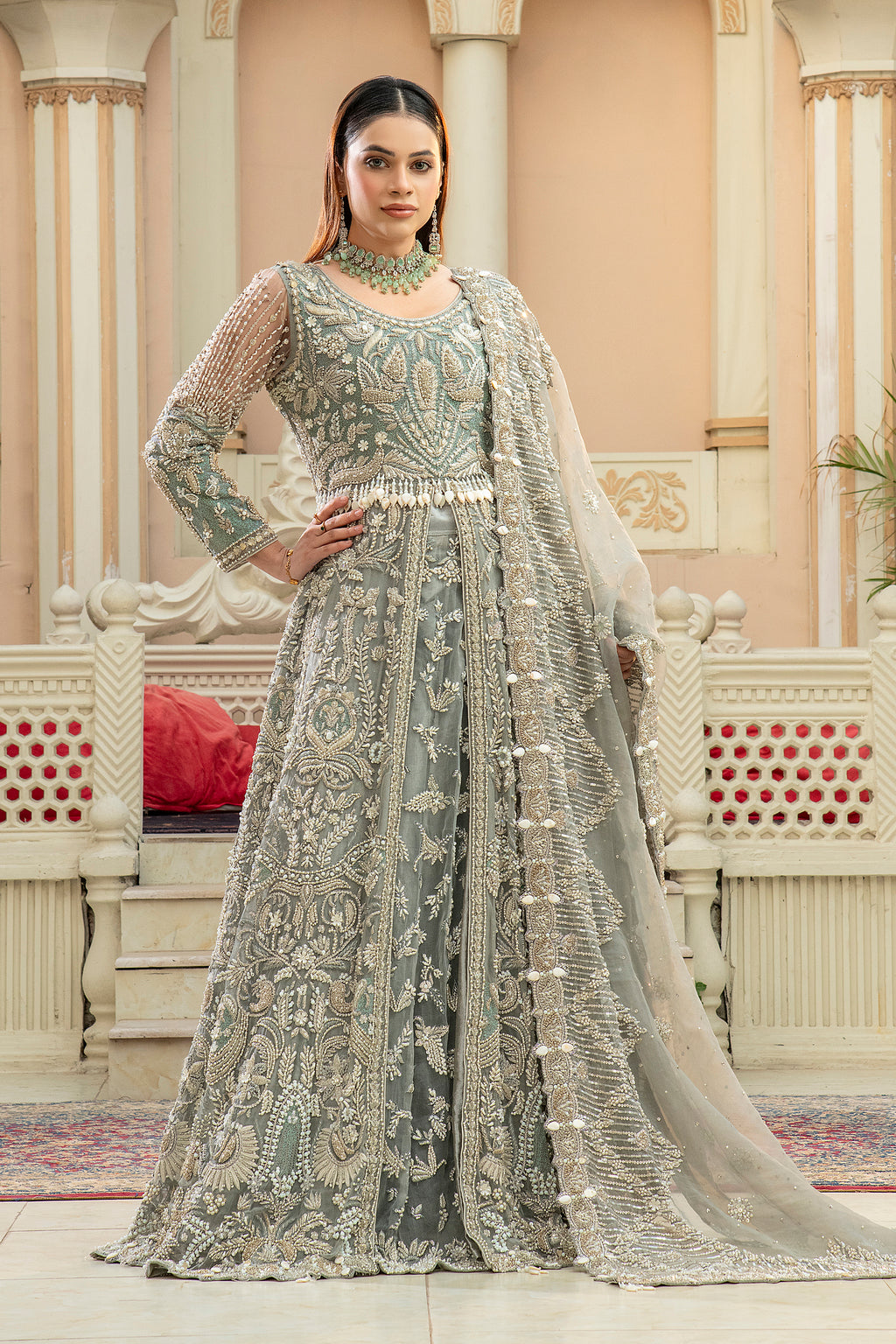 A woman wearing a gray maxi lehnga set with intricate embroidery and jewelry work, standing in a beautifully decorated room.