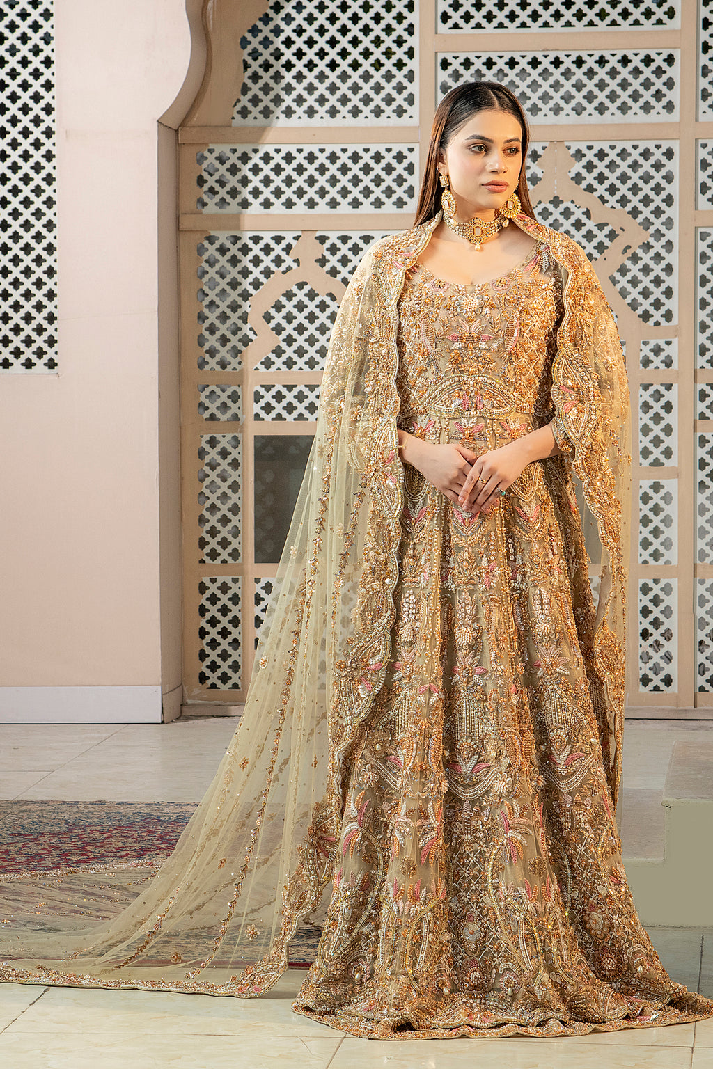 A woman wearing a traditional, fawn-colored maxi dress with embroidery and a dupatta (overlay) featuring an all-over jaal pattern and elaborate embroidery.