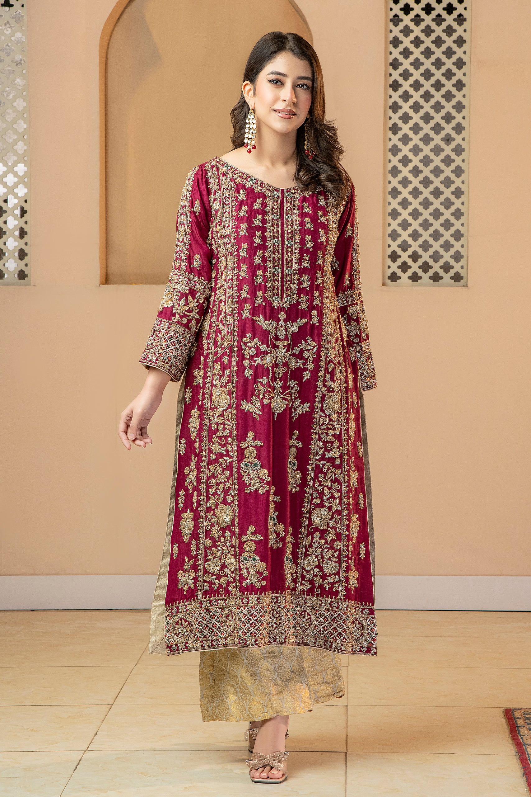 A woman wearing a traditional, vibrant red and gold silk dress with a dupatta, standing in a room with a reflective floor and decorative walls.