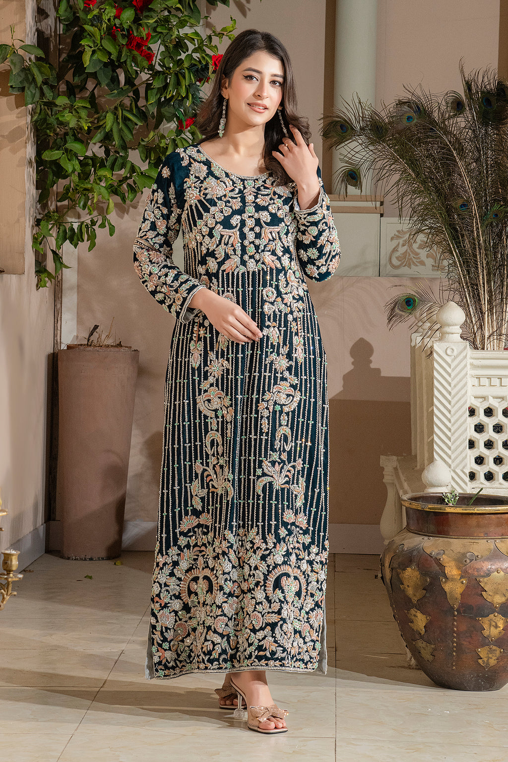 A woman wearing a traditional, embroidered, sea green velvet dress with a dupatta, standing in a room with decorative plants and furniture.