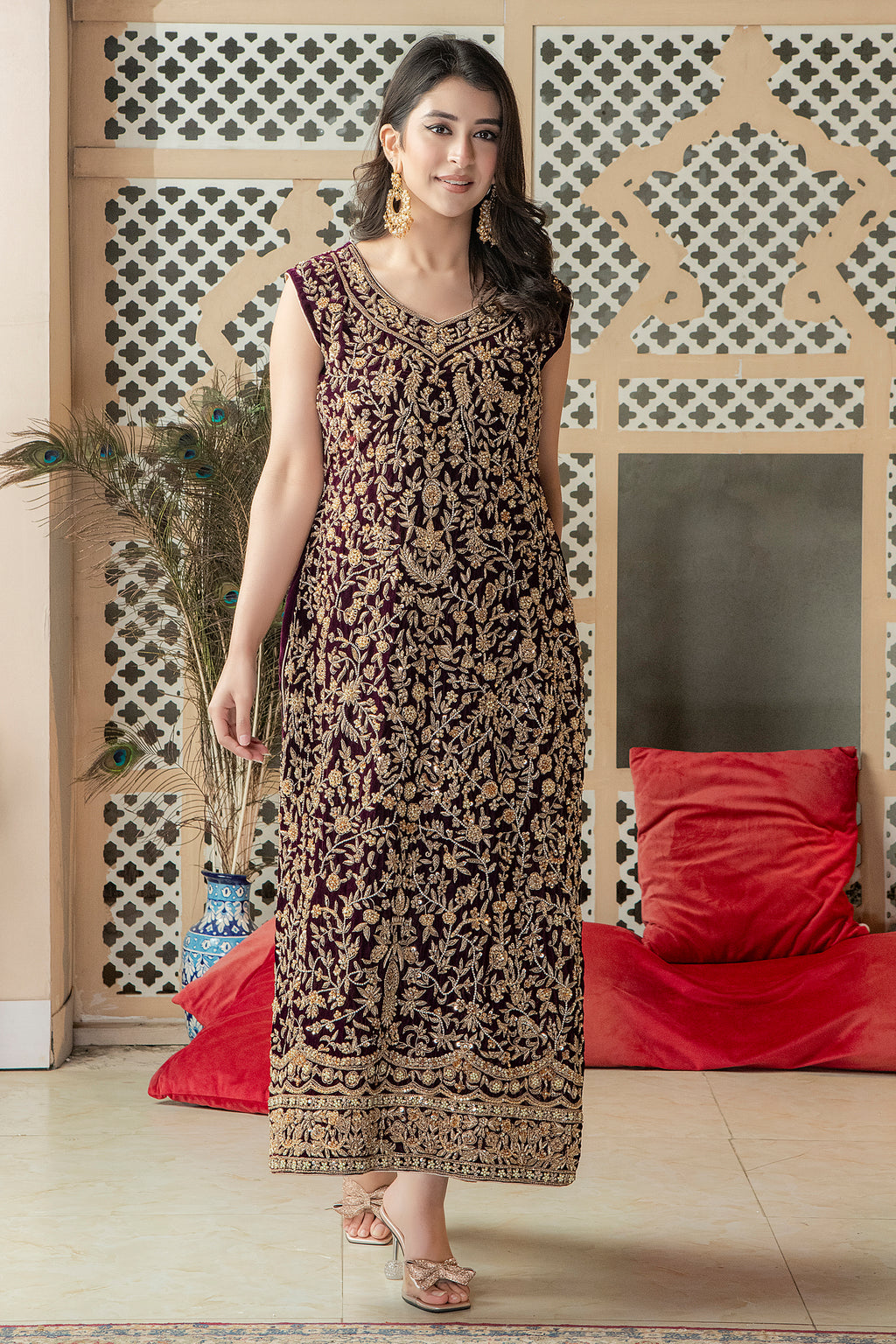 A woman wearing a traditional, embroidered, floral and geometric patterned dress with a dupatta, standing in a room with a red cushion and a decorative wall in the background.