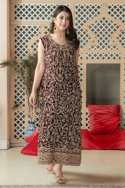 A woman wearing a traditional, embroidered, floral and geometric patterned dress with a dupatta, standing in a room with a red cushion and a decorative wall in the background.