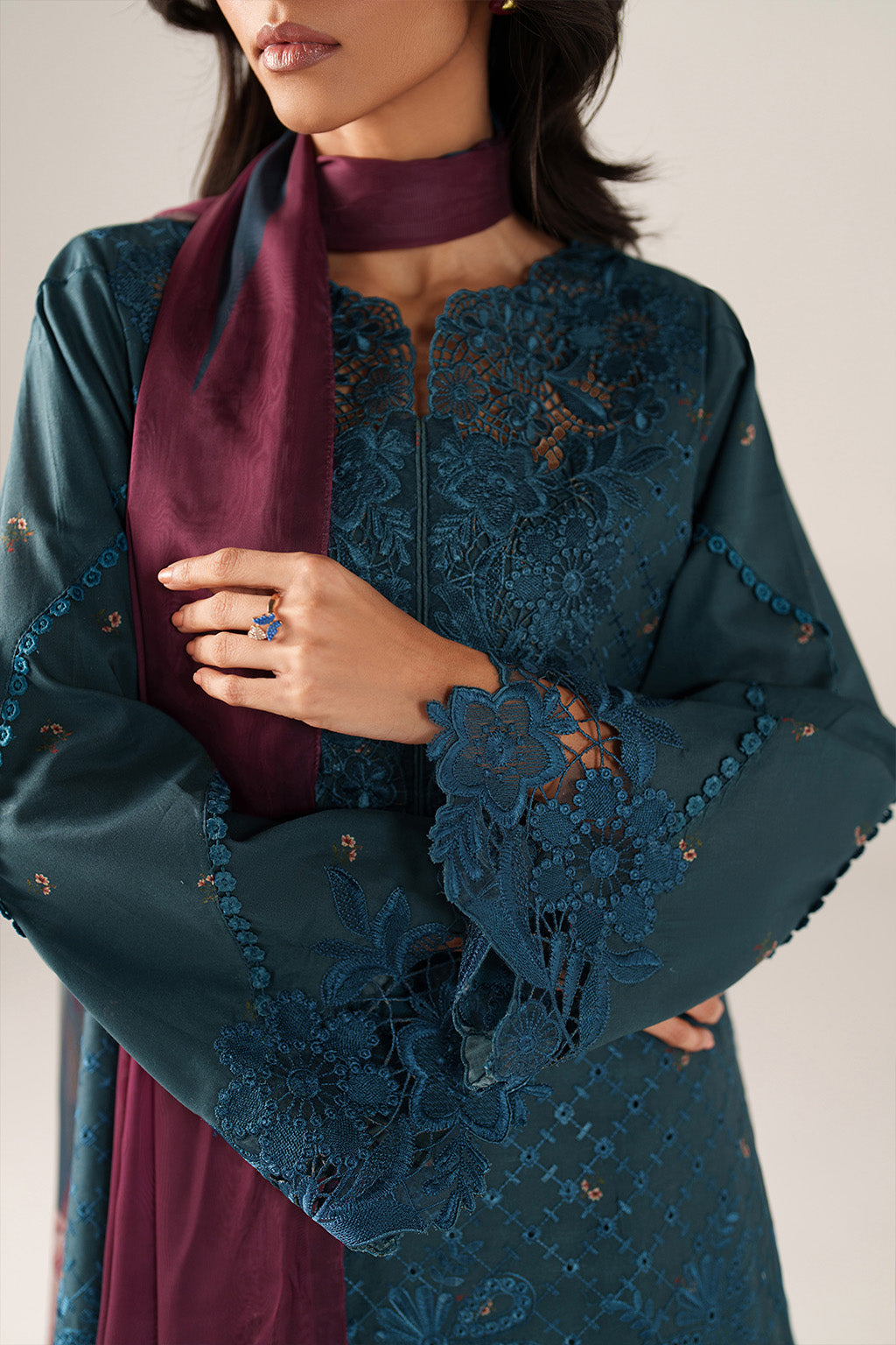 Woman wearing a teal embroidered outfit with a maroon scarf and ring on a neutral background