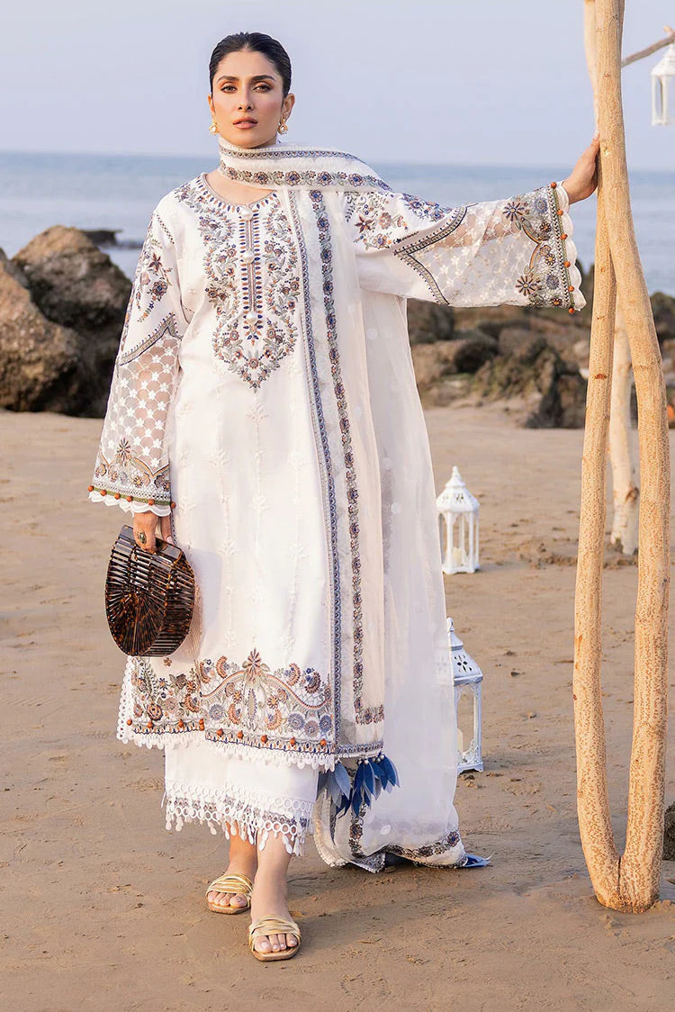 A woman stands on a sandy beach wearing a long white embroidered traditional outfit with beige and brown patterns, holding a small woven bag and leaning against a wooden post.