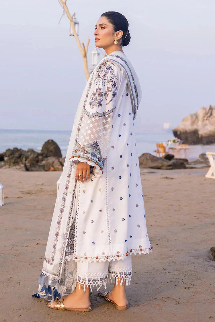A woman stands on a sandy beach wearing a white traditional outfit with blue and silver floral embroidery, a matching dupatta, and wide-leg trousers.