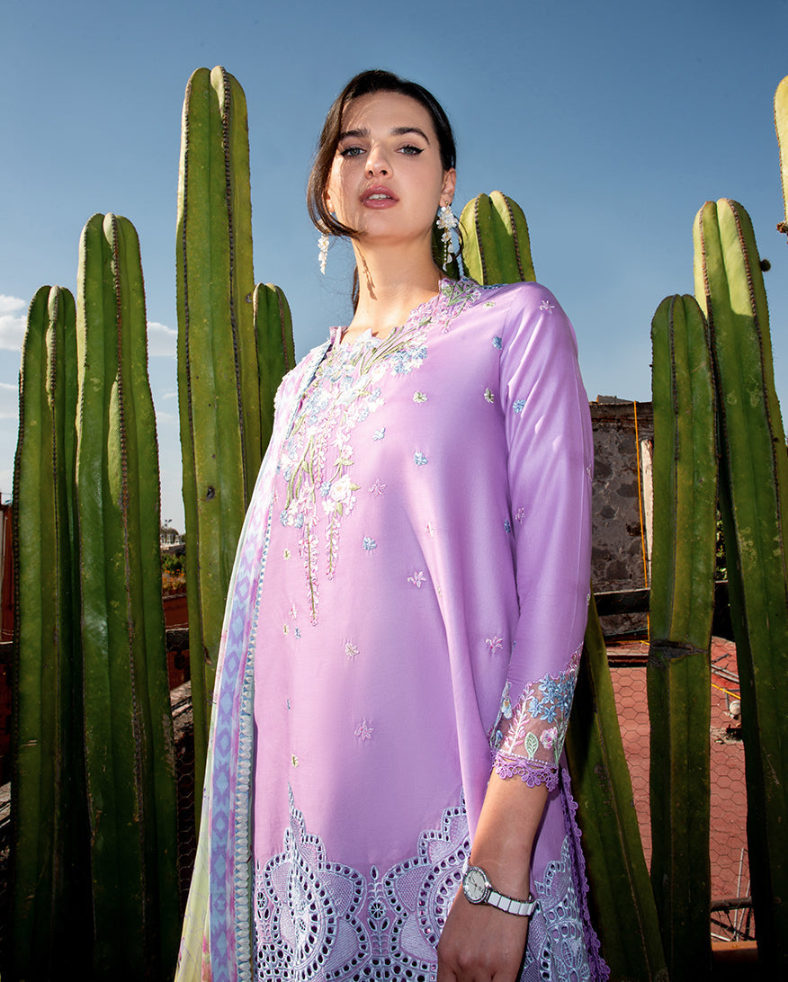 Woman in a light purple traditional outfit standing among cacti with a clear blue sky.