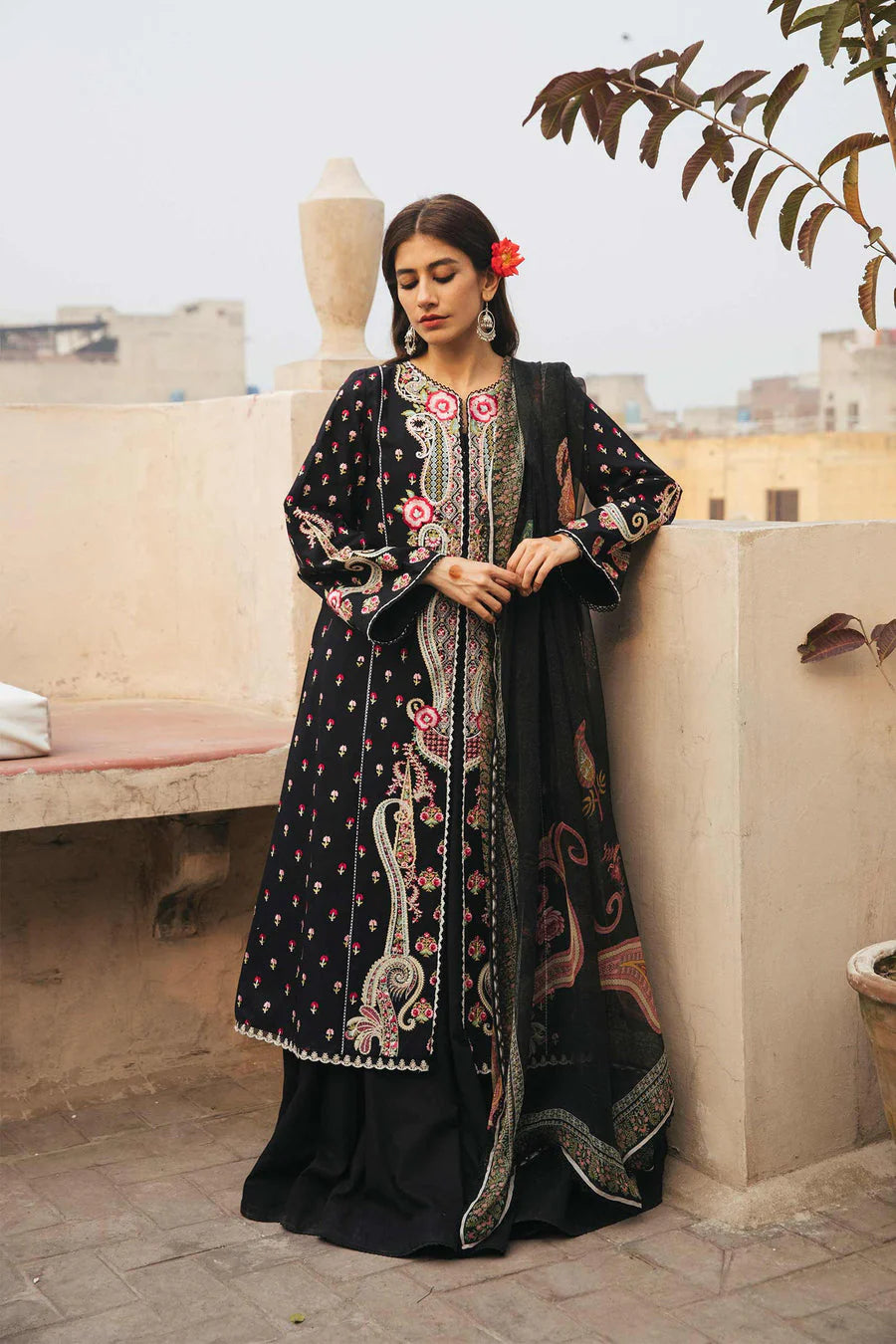 A woman stands on a rooftop terrace wearing a black traditional embroidered outfit with pink and silver floral patterns and a matching dupatta.